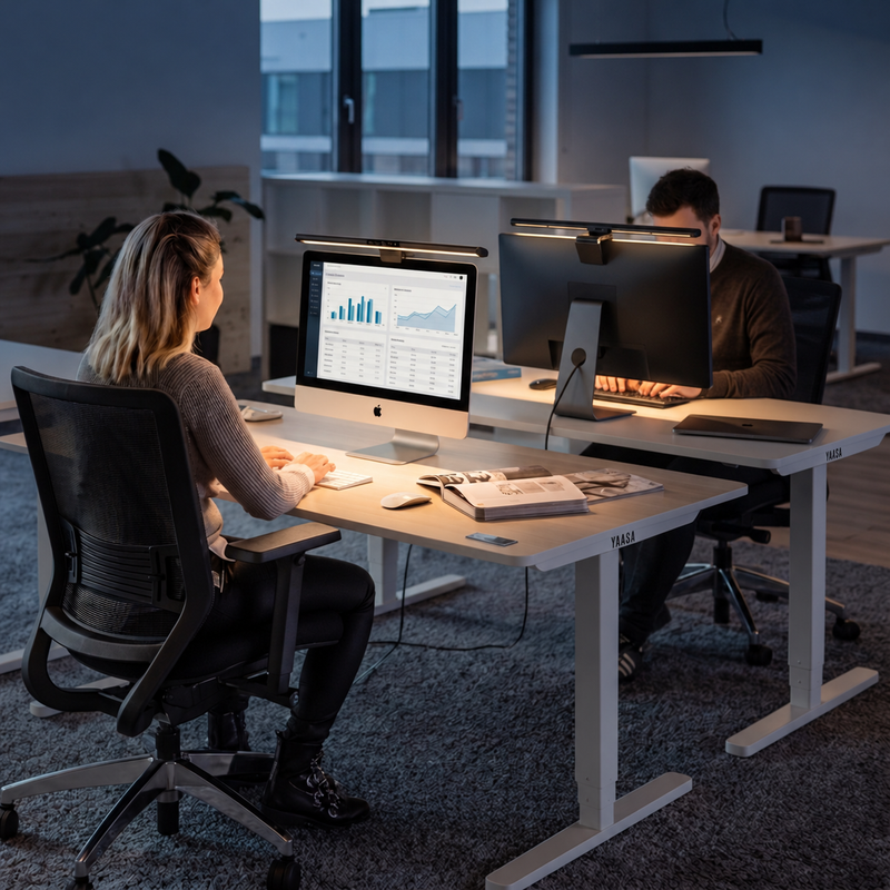 Zwei Personen arbeiten an höhenverstellbaren Schreibtischen mit YAASA Monitor Lampen zur Bildschirmbeleuchtung in einem modernen Büro.