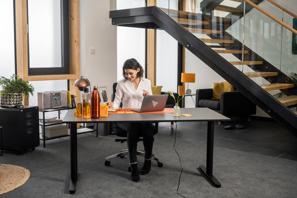 Frau arbeitet im Sitzen an einem schwarzen höhenverstellbaren Schreibtisch in einer Büroumgebung