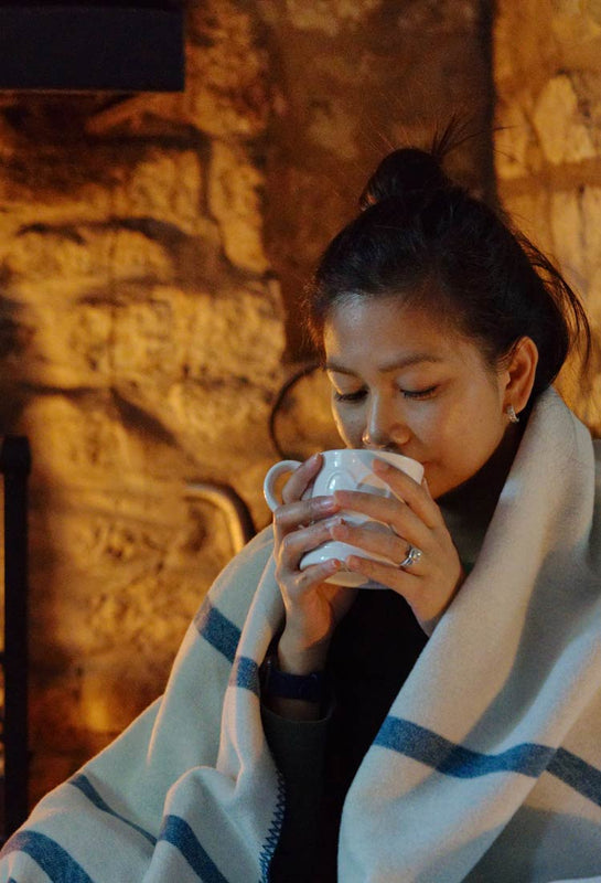 Frau trinkt einen Tee und ist in die Yaasa Elements Blanket eingewickelt.