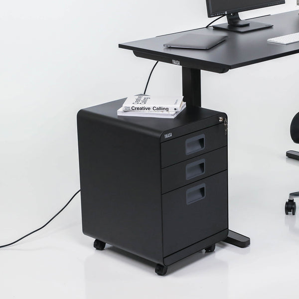 Schwarzer Rollcontainer von Yaasa neben dem elektrisch höhenverstellbaren Schteibtisch Desk Basic in der Farbe Anthrazit.